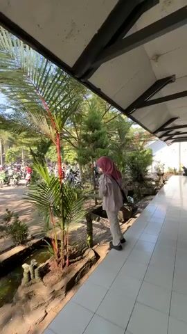 A woman wearing a pink hijab and light-colored clothing walks across a wooden bridge. She is carrying a dark bag over her shoulder. The bridge crosses a small stream. Behind her, a parking lot is visible, filled with motorcycles. Trees and other greenery surround the area, with sunlight filtering through the leaves. The scene appears to be outdoors during the day.