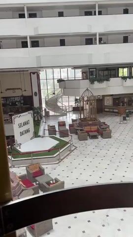 The video shows an empty hotel lobby with a large open space in the center. There are multiple floors visible above the lobby, with balconies overlooking the central area. Various seating arrangements, including wicker furniture with red cushions and low tables, are scattered throughout the space. In the center of the lobby, a raised circular platform is decorated with artificial grass and a sign that reads "SELAMAT DATANG," which translates to "Welcome" in Malay. A spiral staircase is visible in the background, along with a decorative birdcage-like structure with seating inside. The floor is made of white marble with a diamond pattern of small beige tiles. Plants are placed strategically around the lobby, including a tall bamboo plant in the foreground. The lighting appears to be natural light filtering in from windows and interior lighting. There are no people visible in the video.
