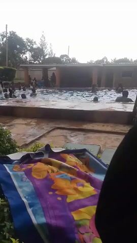 The video shows an outdoor swimming pool filled with people. A man and a young child walk along the poolside holding hands. In the foreground, a colorful blanket is spread out on a raised platform, with some greenery around it. Other individuals are standing and swimming in the pool, and some people are sitting under a covered area near the pool. The weather appears overcast.
