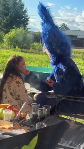 A young girl with long, dark hair is seated outdoors, facing a person in a blue furry costume with a large, pointed headpiece. The costumed person is applying face paint to the girl's cheek with a brush. The girl is wearing pajamas. A table in front of them has plates of fruit, drinks in clear cups, and other items. The background shows trees, grass, and a building. The sky is visible, and it appears to be daytime.