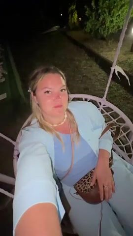 A woman with blonde hair, wearing a light blue outfit and a leopard print purse, is sitting in a pink hanging chair at night. She smiles at the camera, which is held by someone off-screen. The background shows a garden with trees and lights. The chair swings gently back and forth.