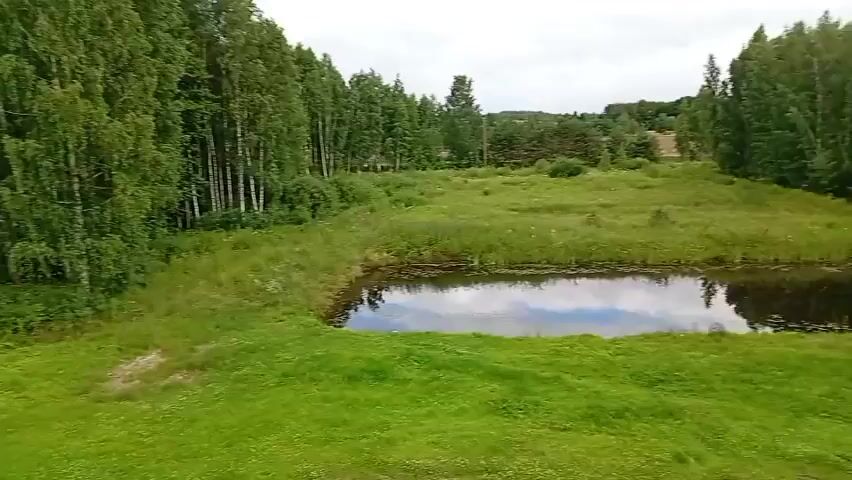 A drone flies over a rural area with a small pond and trees. A doghouse sits near the pond. A small orange car is parked on a dirt path. A brick building is visible on the right side of the frame. The scene is green and lush, suggesting it is summer. The drone moves from left to right, then pans down to show more of the car and the building.