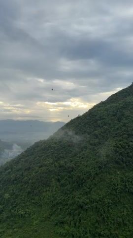The video shows a landscape scene with a steep, green, forested mountain in the foreground. The mountain dominates the right side of the frame. In the distance, several hot air balloons are visible against a cloudy sky with patches of sunlight breaking through. A hazy valley with some structures and possibly agricultural fields is visible below the balloons. The overall lighting suggests it may be early morning or late afternoon.