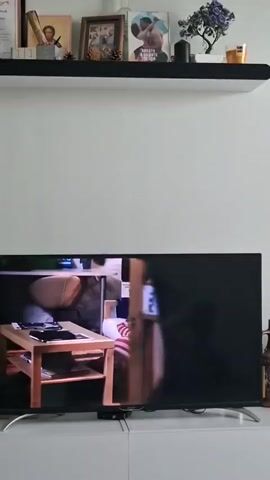 The video shows a TV screen displaying a scene from a movie or TV show. First, a man with a mustache, wearing a black suit jacket over a light blue button-down shirt, looks directly into the camera. Then, the screen shows a woman with long, dark hair, lying face up on a white couch, wearing a red and white striped shirt. The TV is on a white cabinet in front of a white wall. A black shelf above the TV holds various decorative items.