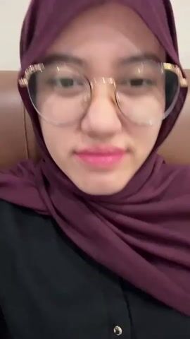 The short video features a close-up of a young woman's face, captured from a selfie perspective. She is wearing a dark purple hijab that drapes around her neck and shoulders, paired with a black collared top, which has a small gold button visible. Her eyes are framed by stylish round eyeglasses with gold-colored frames and decorative accents near the temples. Her lips are adorned with bright pink or fuchsia lipstick.

Throughout the clip, she makes direct eye contact with the camera, offering subtle changes in expression. Initially, her gaze is direct and neutral. She then briefly lowers her eyes and tilts her head slightly, revealing a hint of a smile. This movement is repeated with a more pronounced tilt, and she glances off-camera before returning her gaze. The background is a blurred warm brown, suggesting upholstered seating like a chair or couch in an indoor setting. The overall mood conveyed is calm and slightly playful. There is no visible text in the video.