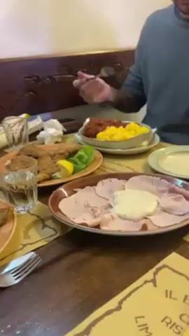 A wooden table holds several dishes of food, including a plate of thinly sliced meat with a creamy sauce, a plate of breaded cutlets with lemon and lettuce, and a dish of polenta and a red sauce. A person is seated at the table, holding a fork and reaching towards the food.  The table also has glasses of water, silverware, napkins, and a menu or placemat with text. The background features a carved wooden design on a wall.