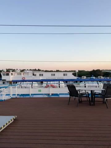 The video captures a close-up, side-view of a white cruise ship moving through calm water. The ship has a blue railing running along its side and a brown deck in the foreground. A table with four chairs is set up on the deck. In the background, trees line the shore, and the sky is a clear, pale blue with a hint of pink as if at sunset. The ship's name, "ALEXANDER PUSHKIN," is visible on its side. Life rafts and life preservers are also apparent on the ship's deck. The video is shot at eye-level from the deck of the ship itself, giving a sense of being on board.
