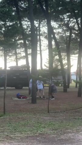 The video shows three people standing in a wooded area, with a black van and a trailer visible in the background. The sun is shining through the trees, creating a hazy, late afternoon or early evening light. The ground is covered with dry grass and pine needles. One person wears a white top, shorts, and a hat. The other two people wear darker clothing. There are backpacks and other items on the ground. The people appear to be talking, but their conversation is not clear.
