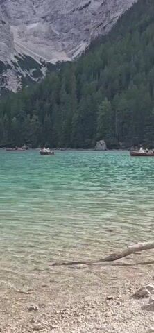 A serene, wide shot captures a turquoise lake with visible pebbles and rocks beneath the surface. The lake is dotted with several small, wooden rowboats, some of which contain passengers. In the distance, a dense, dark green forest lines the shore, with a massive, rocky mountain rising majestically behind it, its peaks shrouded in a light haze. The sky above is overcast with light clouds. A single, fallen branch rests on the sandy shore in the foreground.