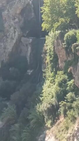 The video captures a natural scene with a waterfall cascading down a rocky cliff face. Lush green vegetation, including trees and bushes, covers parts of the cliff and the surrounding area. The lighting suggests it is daytime, with sunlight illuminating the upper sections of the trees and the cliff. The viewpoint is from a distance, looking up at the waterfall and the landscape. There are no discernible people or man-made objects in the frame, emphasizing the natural beauty of the environment. The movement in the video is limited to the flow of the waterfall and potentially subtle shifts in the foliage due to wind.