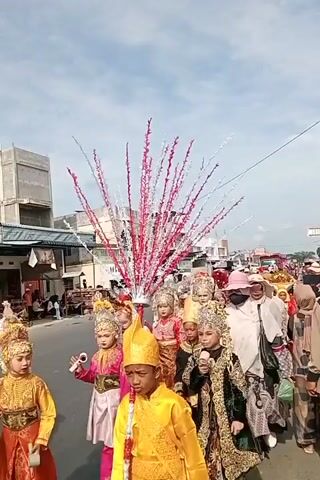 The video shows a procession on a street, possibly a parade or a cultural event. Several individuals are walking, some in traditional attire with elaborate headdresses and costumes. In the foreground, a young person in a yellow costume with a golden headdress is visible. Behind them, a group of children and adults are walking, many wearing colorful and ornate outfits. Some are wearing masks. The environment appears to be a town or city street with buildings on either side, some with shops. The sky is visible and appears to be partly cloudy. The procession is moving forward, and onlookers are present along the sides of the street.