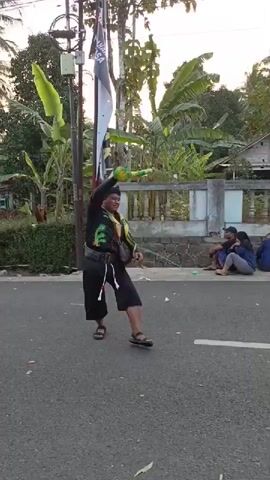 A man in black clothing with a green and yellow sash performs a martial arts routine on an asphalt road. He is holding a long, flexible green and yellow object, possibly a whip or a rope. A flag with a black and white design is attached to a pole behind him. In the background, several people are sitting on the side of the road, and there are trees and houses. The sun appears to be setting, casting long shadows.