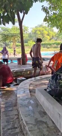 The video depicts a scene at a public swimming pool on a sunny day. Several individuals are visible, some in the water and others on the poolside. In the foreground, a person wearing a red shirt is sitting on the edge of the pool with their feet in the water, while another person in a black headscarf is also seated nearby, with their feet submerged. To the right, a young person with dark curly hair, wearing a black headscarf, is sitting by the pool's edge. In the background, a person in a red shirt and black shorts is walking away from the camera, towards the pool. There are other individuals in the pool, including children. The pool area is surrounded by concrete walkways and some vegetation, with trees visible in the background. The overall environment appears to be a recreational outdoor space.
