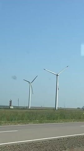 The video shows a view from inside a vehicle, likely a car, traveling on a road. The foreground shows a section of the road with white lane markings. Beyond the road is a grassy field with some scattered yellow flowers. Two large white wind turbines are prominently visible in the middle ground, with their blades extended. In the far distance, more wind turbines can be seen spread across the horizon, suggesting a wind farm. The sky is clear and blue. The video also captures parts of the vehicle's interior, including the windshield, side mirror, and a section of the car's roof or pillar, indicating the perspective of the camera is from a passenger or driver. There are no people visible in the video.