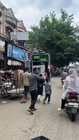 The video opens on a street scene with people and vehicles. In the foreground, a person on a motorcycle is visible from the chest down. To the left, a man in a checkered shirt and dark pants walks towards the camera, followed by a cart. A woman in a light blue hijab and dress, carrying a large pink cotton candy, walks next to a young boy in a patterned shirt. Behind them, a white truck with green speaker boxes on top is parked. A person in a black shirt is interacting with the back of the truck. Further down the street, other motorcycles and a black SUV are visible. The street is lined with buildings and trees, suggesting an outdoor marketplace or commercial area. The ground is paved, and there are some trash items scattered on it. The lighting suggests it is daytime with overcast skies. The camera pans slightly to the right, following the motorcycle in the foreground as it moves down the street. The scene is bustling with activity, indicating a public gathering or event.