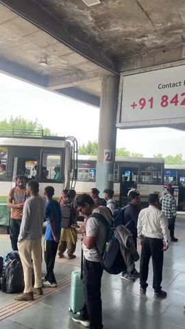 The video shows people standing and waiting in line to board green and white buses at a bus station. Many individuals have luggage such as suitcases and backpacks with them. Some people are looking at their phones or talking to each other. The bus station has signs with phone numbers on them. It is under a covered area with a gray concrete ceiling. The sky visible outside is overcast.