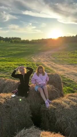 Two women are seen sitting on large hay bales in a rural field at sunset. The sun is low on the horizon, casting a warm glow across the scene. The sky is partly cloudy. In the foreground, there are several large, round hay bales. A green field extends behind the hay bales, leading to a line of trees in the distance.

The woman on the left is wearing a black long-sleeved jumpsuit and is in the process of adjusting her hair, with both hands raised to her head. A phone or small object is placed on the hay bale next to her. The woman on the right is wearing a light pink long-sleeved top and light-colored shorts, and is looking down at her lap. Both women are wearing sneakers. They appear to be casually posing or resting in the field.

Overlaid text in Russian is visible: "Сейчас подожди, Серёж, меня тоже надо будет." (Wait a minute, Seryozha, I also need to be [in the shot].), "Жар" (Heat), and "Только на мой телефон" (Only on my phone). The location context provided is Стеклянный, Russia.