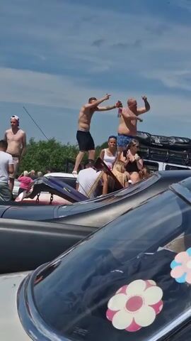 The video captures an outdoor gathering under a partly cloudy sky, with a focus on people interacting around cars. In the foreground, the shiny hood of a car with floral stickers is visible. Behind it, a dark-colored car is parked, and further back, another light-colored vehicle is partially obscured.

The central action involves several individuals. Two men are standing on top of the light-colored car, with one holding a bottle and gesturing. Another man is standing nearby on the car's roof, also with a bottle. To their right, two women are seated on the edge of the car. Several other people are visible in the background, some standing near the cars, and one person in a pink shirt is seen walking away from the group. The overall atmosphere appears to be casual and social, likely at an event or informal gathering.