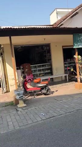A red scooter with an orange seat is parked on a brick sidewalk in front of a storefront. The storefront has an open entrance and display shelves visible inside. A person wearing a floral dress is standing inside the store. A white bench is located outside the store to the right of the entrance. The scene is bathed in bright sunlight, casting shadows on the ground.