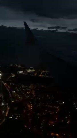 The video shows the wing of an airplane at night, with the lights of a city visible below. The sky is dark, with clouds. The wing of the plane is gray, with the Ryanair logo visible. The city lights are orange and yellow, creating a speckled pattern. The overall scene is dark and atmospheric.