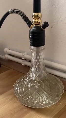This is a close-up, medium shot of a hookah setup, filmed from a slightly low angle. The hookah has a clear, diamond-patterned glass base filled with water. The stem of the hookah is black with gold accents and a black hose with a gray ribbed sleeve is attached. The hookah is positioned on a wooden floor against a white wall. Two white pipes or conduits are visible along the bottom of the wall behind the hookah. The video captures the sound of water bubbling, indicating that the hookah is in use, with air being drawn through the water.
