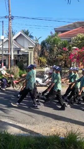 A group of young men in matching green patterned shirts, dark pants, and blue caps are marching in formation on a paved street. They are performing a synchronized routine, with some raising their arms to the side and others extending them forward. The sun is shining, and the sky is clear and blue. Houses and trees line the street, and a motorcycle is visible in the background. Some of the marchers are wearing white gloves. The scene appears to be a parade or a drill practice.