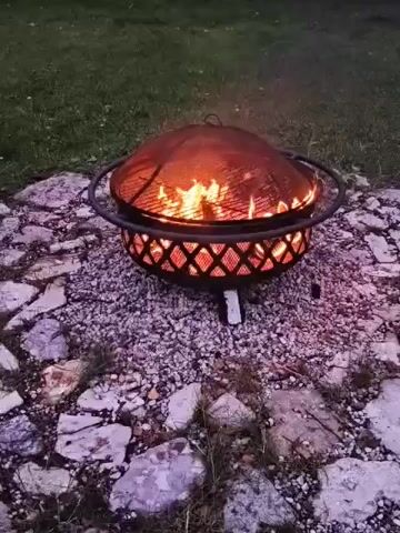 The video shows a fire pit in an outdoor setting. The fire pit is round with a decorative metal lattice around the base and a mesh lid. Flames are visible inside the fire pit. The fire pit is placed on a bed of gravel and surrounded by irregular stones and grass. The background is a lawn with trees and foliage, suggesting it is a yard or garden. The lighting indicates it is dusk or evening. There are no people visible in the video. The camera is mostly stationary, with slight movements, focusing on the fire pit.