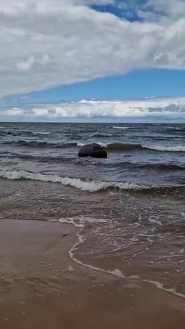 A large, dark rock sits in the choppy waters of a lake or sea. Waves crash against the rock, sending white spray into the air. The shoreline is visible in the foreground, with sand and small waves lapping at the shore. The sky above is cloudy with patches of blue.