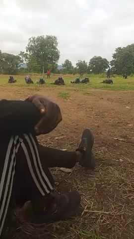 The video shows a group of people in an outdoor setting. In the foreground, a person is sitting on the ground with their legs extended, wearing black track pants with white stripes and black athletic shoes. Their hands are clasped. In the background, several other people, dressed in dark clothing and possibly military uniforms, are lying on the ground in a prone position, performing some form of exercise or training. The environment appears to be a field with dry grass and dirt, surrounded by trees and a slightly cloudy sky. One person in the background is standing and appears to be observing or instructing the group. The overall scene suggests an outdoor training or physical activity session.