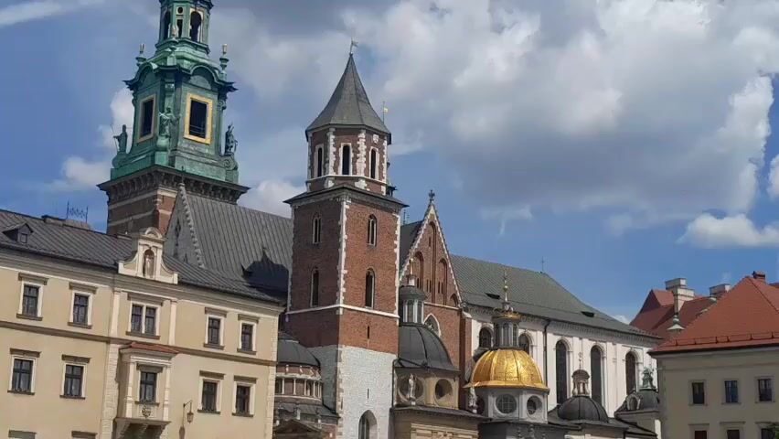 The video displays a close-up view of the Wawel Cathedral in Krakow, Poland, under a partly cloudy sky. The camera is positioned at a low angle, emphasizing the imposing architecture of the cathedral and adjacent buildings. The foreground shows a section of a light-colored stone building with multiple windows. To the right, the Wawel Cathedral is visible, featuring a prominent brick tower with white accents and a grey, conical roof topped with a spire. Adjacent to this tower, a larger, taller green-copper clad tower with ornate architectural details and a clock face is visible. Several smaller domes, some gilded, are also part of the cathedral complex. The buildings are constructed with a combination of stone and brick, with tiled roofs. The sky is a bright blue with scattered, fluffy white clouds. No people or specific actions are discernible in this static shot.