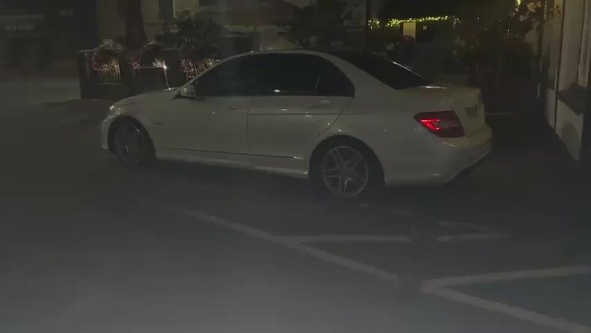 A white Mercedes-Benz C-Class sedan is parked on a street at night. The car is facing left, and its rear is visible. The car is parked in a marked parking space. There is some fog or mist on the ground around the car. In the background, there are buildings and some trees.