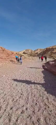 The video shows a group of people walking on a flat, sandy, light brown landscape. There are hills on either side of them with varied shades of brown and some red. The sky is clear and blue.  The people in the group vary in dress, one individual carrying a bright blue parasol. The shadow of the person holding the camera can be seen in the foreground.