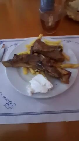 The media file shows a close-up, slightly blurry, overhead shot of a meal on a table. The meal consists of ribs and french fries served on a white plate, with a dollop of white sauce or cream on the side. A fork is visible on the right side of the plate. The table is covered with a placemat that has a blue and white pattern. In the background, out of focus, is a glass with a dark liquid, a blue can or bottle, and a basket with bread. Further in the background, on an orange-colored wall, are framed pictures. One picture is a dark rectangular frame with text and an illustration that appears to be a woman playing golf. The text under the illustration reads "British Ladies Open 1929". Another frame is partially visible to the right. The lighting suggests it is daytime, possibly indoors at a restaurant. The overall impression is of a casual meal.