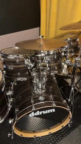 The video shows a drum set in a room. The drum set is dark colored with chrome hardware. There are several cymbals, a bass drum, a snare drum, and at least two tom-toms. The background is a yellow curtain. The floor is a black mat with a diamond pattern.