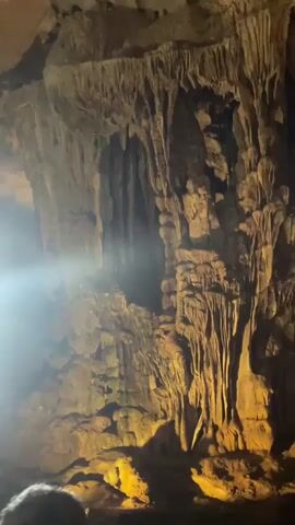 The video pans across a dimly lit cave, showcasing impressive stalactites and stalagmites illuminated by artificial lighting. A bright shaft of light beams down from an opening in the cave ceiling, creating a dramatic effect and highlighting dust particles in the air. In the foreground, the backs of several people are visible, suggesting they are part of a tour group exploring the cave. The overall atmosphere is one of awe and natural wonder.