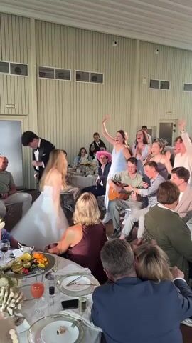 A wedding reception is in full swing. A groom in a tuxedo and a bride in a white wedding dress are dancing in the center of the room. A woman in a light blue dress is singing and clapping along with the music, while a group of people are gathered around a man playing a guitar. Guests are seated at tables, some are clapping and cheering. The room is decorated with pale wood paneling and white walls. A table in the foreground is laden with food and drinks.