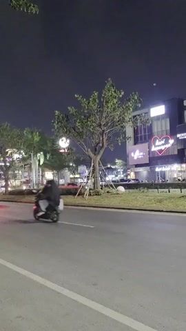 The video shows a street at night. A tree stands in the middle of a grassy median. Several cars are visible on the road. Buildings with illuminated signs are on the right side of the street. One sign displays a large heart shape. A person is riding a scooter in the left lane.