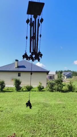 A black wind chime with horse-shaped ornaments hangs from a square top against a bright blue sky with sparse white clouds. The wind chime is in the foreground, with a blurred green lawn and a light-colored house with a dark roof in the background. Trees and other houses are visible behind the main house. The wind chime is gently swaying, causing the horse ornaments to move. The sunlight indicates it is daytime.