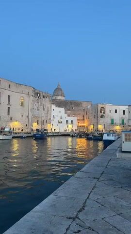 The video shows a waterfront scene at dusk. The water in the foreground is dark and reflects the lights from the buildings and boats on the other side of the harbor. The buildings are made of light-colored stone and have multiple stories. Some of the buildings are lit from within, casting a warm glow on the water. Several boats are docked along the waterfront, including a white motorboat with a yellow stripe. In the background, a large, domed building is visible, as well as the silhouette of a castle-like structure. The sky is a deep blue, transitioning to a lighter shade near the horizon, suggesting the end of daylight. There are lights from streetlamps and buildings illuminating the area. The video pans slightly to the right, revealing more of the harbor and the buildings lining it. There are a few people visible on the docks, but they are small and indistinct.