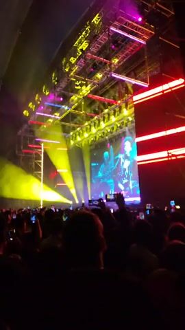 The video shows a concert scene from the perspective of someone in the audience. The crowd is dense, with many people holding up their phones, seemingly recording the performance. On stage, there is a large screen displaying a video of a musician. The lighting is bright and colorful, with beams shining down on the stage. The concert appears to be held inside a large tent-like structure, as the roof is visible in some parts of the video. The energy is high, with people presumably enjoying the music and show.
