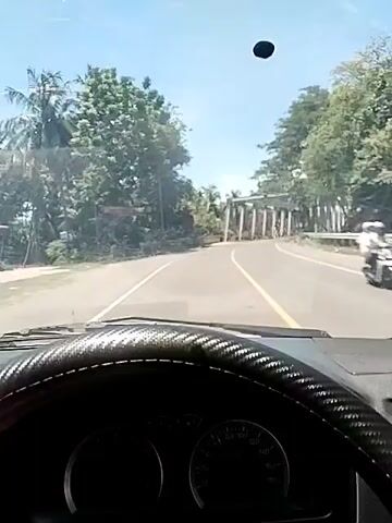 From a car’s dashboard perspective, a driver approaches and proceeds through a metal truss bridge. The day is sunny, with palm trees visible in the background. Cars and motorcycles pass in the opposite direction. The steering wheel has a black and red cover. A black spot is present on the windshield.