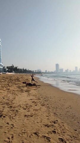 On a sandy beach, two individuals are engaged in physical exercises. The first person is performing push-ups. The second person is doing lunges. Other people are present on the beach, some visible near the shoreline. The ocean's waves gently roll in, reflecting the hazy sunlight. The backdrop of the scene consists of a cityscape, defined by high-rise buildings silhouetted against the horizon. A few trees and street lights are visible along the beach's edge. The sky is overcast.
