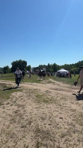 The video shows an outdoor event on a clear, sunny day. In the foreground, a person walks away from the camera, carrying a bag. To the left, another person walks towards the camera, carrying a folded chair. In the background, there are several tents, a stage with people on it, and a crowd of people gathered. The ground is covered in dirt and grass. The overall atmosphere appears to be festive.