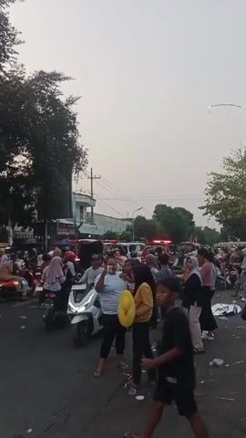 A busy street scene captures a crowd of people and motorcycles. The sky is overcast. A white motorcycle is prominently featured in the foreground. Many people are dressed in casual clothing, with some women wearing hijabs. Several red vehicles with flashing lights, possibly emergency vehicles, are visible in the background, suggesting a possible incident or event. The street appears to be congested with both people and vehicles, indicating a crowded public space. The overall impression is one of a bustling urban environment with a significant gathering of individuals.