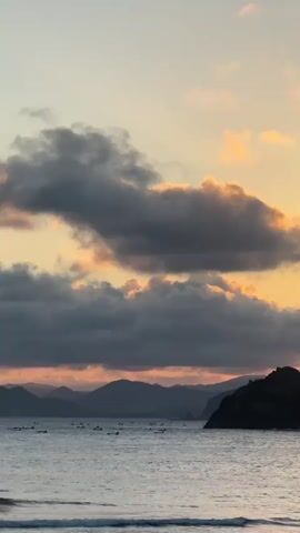 This is a vertical video capturing a serene ocean view at sunset in Selongbelanak, Indonesia. The scene is dominated by a dramatic sky filled with a mix of dark, heavy clouds and lighter, illuminated clouds tinged with orange and yellow hues, suggesting the sun is setting or has just set.

The ocean in the foreground is relatively calm, with gentle waves lapping at the shore. A dark, rocky outcrop juts out into the water on the right side of the frame, providing a stark contrast to the softer elements of the sky and sea. In the distance, a series of silhouetted mountains form the backdrop, their peaks softened by the atmospheric haze.

Scattered across the water, particularly in the middle ground and stretching towards the distant shore, are numerous small boats, possibly fishing vessels or surfboards. This suggests a coastal community activity, perhaps related to fishing or surfing, is taking place.

The overall mood is peaceful and tranquil, with the warm colors of the sunset creating a sense of calm and beauty. The time of day is clearly evening, as indicated by the sunset and the subdued lighting. There are no visible text elements in the video.