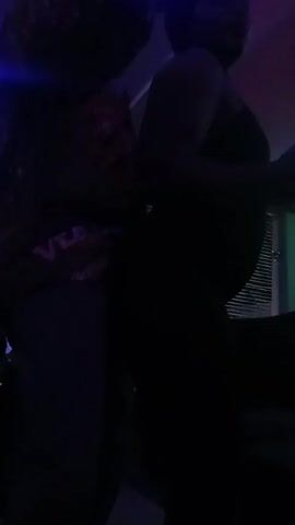 A dimly lit video shows two figures, one partially visible, dancing to music. The figure on the left is wearing a dark shirt with a "VERSACE" print on the back. The environment seems to be indoors, with slatted blinds visible in the background. There's also a blue light adding to the ambiance.