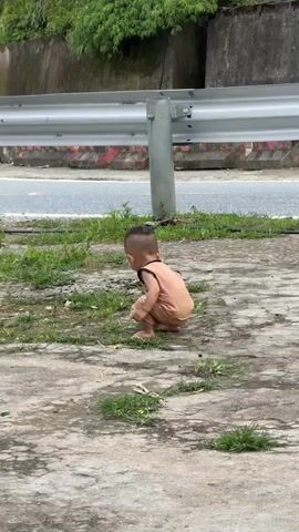 A medium shot shows a toddler in a brown sleeveless top and shorts, barefoot, squatting on a concrete and grass surface. They hold a brown object in their left hand, which they drop. They then stand up and turn slightly to their right. In the background, a white car drives past a metal barrier on a road. There are green trees and a concrete wall behind the road.