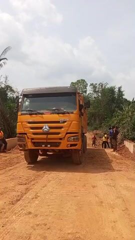 A large yellow HOWO SINOTRUK 37 dump truck is slowly reversing on a wide, reddish-brown dirt road under a bright, cloudy sky. Several individuals, mostly men, are visible in the scene. One person is seated on a low concrete wall to the left of the truck, wearing an orange safety vest, while others stand nearby or further down the road. Dense green vegetation and trees frame both sides of the road and extend into the background. The area appears to be a construction site, possibly for road development or a culvert/bridge, given the presence of excavated dirt piles and the concrete structures. The time of day is clearly daytime, with ample sunlight. The setting is likely rural or semi-rural, consistent with Aloko, Nigeria.