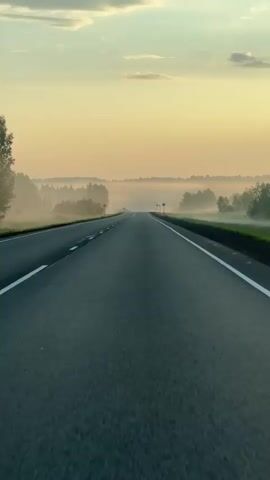 A video shows a drone's eye view of an empty asphalt road stretching into the distance. The road is lined with white dashed and solid lines. On either side of the road are trees shrouded in a light mist, and rolling hills are visible in the background under a pale, hazy sky with scattered clouds. The sun appears to be setting or rising, casting a warm, golden light on the scene. There are no people or vehicles visible. The overall atmosphere is serene and tranquil.