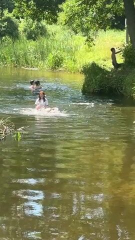 A group of children are enjoying a summer day swimming and playing in a shallow river surrounded by lush green vegetation and trees. The sun is shining brightly, reflecting off the water's surface, creating a dappled effect. The scene suggests a warm and playful atmosphere, with the children clearly having a fun time. 

In the foreground, a young girl with light brown hair, wearing a white shirt, splashes in the water. Behind her, two other children are visible in the river. Further back, a person is about to jump from a grassy bank into the water, their body in mid-air. The river is wide and clear, with the natural beauty of the surrounding landscape adding to the serene and idyllic setting. The time of day appears to be midday, judging by the bright sunlight and the long shadows cast by the trees. The overall mood is one of carefree joy and summer recreation in a natural environment.